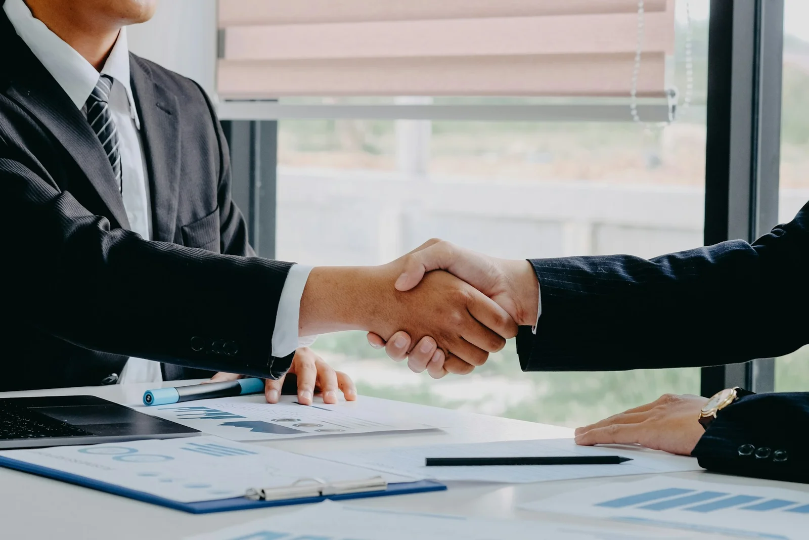 Two people shaking hands over a desk with charts and a laptop, sealing agreed billing terms for a consulting engagement